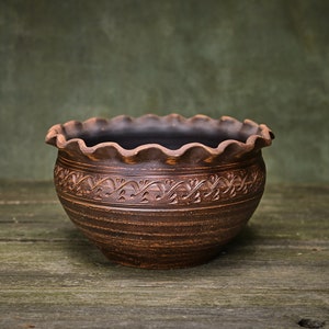 May include: A brown ceramic pot with a scalloped rim and intricate carvings. The pot is sitting on a rustic wooden surface.