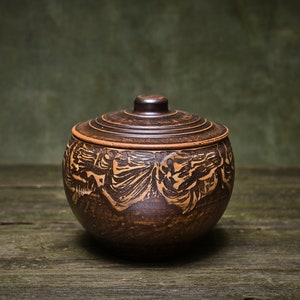 May include: A brown ceramic jar with a lid, featuring intricate carved designs. The jar is sitting on a rustic wooden surface.