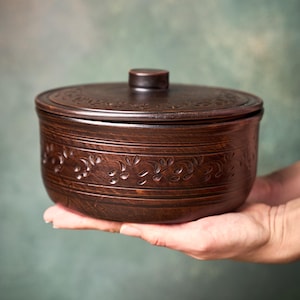 May include: A brown ceramic bowl with a lid, held in a person's hand. The bowl features a floral design around the middle and on the lid. The bowl is round and has a small knob on the lid.