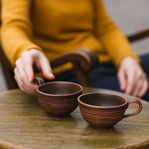Può includere: Due tazze da tè in ceramica marrone con manici sono su un tavolo rotondo di legno. Le tazze hanno un design strutturato e scanalato. Il tavolo è all'aperto e una persona con un maglione giallo è sullo sfondo.