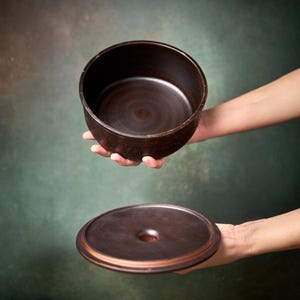 May include: A dark brown ceramic bowl with a smooth interior and a matching circular lid featuring a central circular opening. The bowl has a deep, cylindrical shape with a slightly textured exterior surface and a consistent dark brown finish.