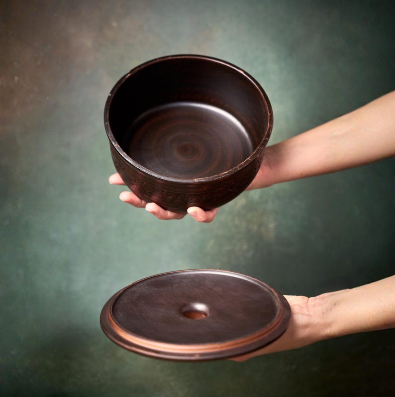 May include: A dark brown ceramic bowl with a smooth interior and a matching circular lid featuring a central circular opening. The bowl has a deep, cylindrical shape with a slightly textured exterior surface and a consistent dark brown finish.