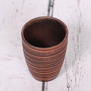 May include: A cylindrical brown clay cup featuring a series of horizontal, incised grooves circling the exterior. The vessel has a smooth, open top rim and a tapered base, showcasing the natural, earthy texture and deep reddish-brown color of the material.