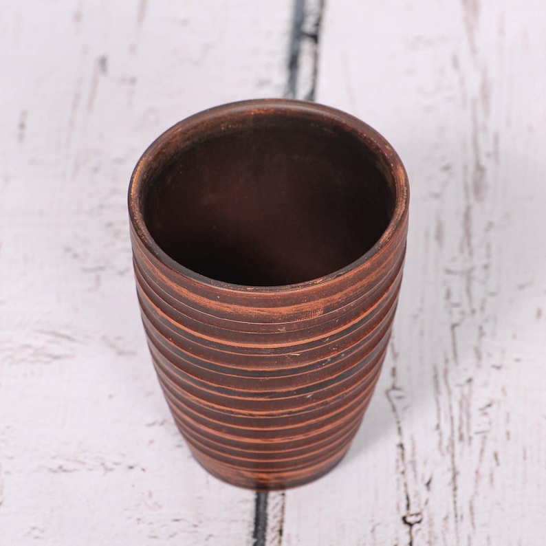 May include: A cylindrical brown clay cup featuring a series of horizontal, incised grooves circling the exterior. The vessel has a smooth, open top rim and a tapered base, showcasing the natural, earthy texture and deep reddish-brown color of the material.