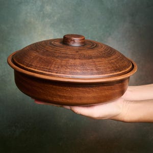 May include: A round, brown clay casserole dish with a matching lid. The exterior of the pot and the lid feature visible horizontal circular grooves and a smooth, earthy texture. The lid has a small, centered knob handle on the top surface.