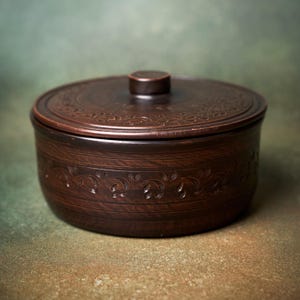 May include: A round, dark brown clay lidded container featuring a smooth finish and a central knob on the lid. The exterior of the vessel and the lid are decorated with a repeating, carved vine and leaf pattern that encircles the entire piece.