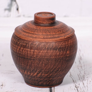 Peut inclure: Un pot en terre cuite brun avec un couvercle. Le pot a une surface rugueuse et texturée.