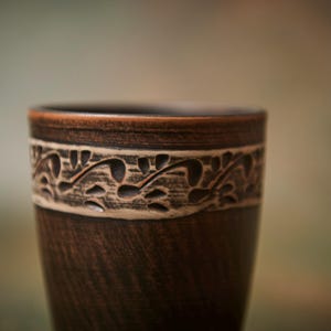 May include: A handcrafted, dark brown ceramic cup with a decorative band featuring a carved floral design. The cup has a smooth, polished finish and a slightly flared rim. The cup is approximately 5 inches tall.