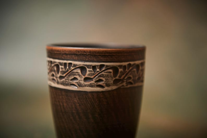 May include: A handcrafted, dark brown ceramic cup with a decorative band featuring a carved floral design. The cup has a smooth, polished finish and a slightly flared rim. The cup is approximately 5 inches tall.
