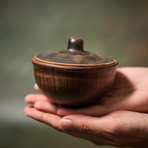 May include: A small, round, dark brown clay bowl with a matching lid. The bowl features a textured, earthy surface with visible horizontal striations and a smooth, rounded knob handle on the center of the lid. The material appears to be unglazed.