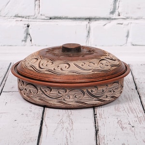 May include: A brown ceramic casserole dish with a lid. The dish has a decorative design of swirling lines and is sitting on a white wooden surface.