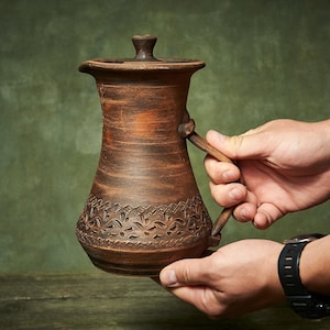 Könnte beinhalten: Ein brauner Tonkrug mit Deckel und Henkel. Der Krug hat einen breiten Boden, der sich nach oben hin verjüngt, und ist im unteren Bereich mit einem geschnitzten geometrischen und floralen Muster verziert. Die Oberfläche ist matt und texturiert.