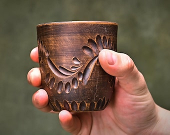 May include: A brown clay cup featuring a hand-carved floral pattern with deep, textured grooves. The vessel has a cylindrical shape with a slightly tapered base and a dark, earthy finish, showcasing visible artisan craftsmanship and rustic details.