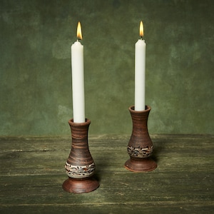 May include: Two lit white candles in brown ceramic candle holders with floral designs. The candle holders are on a weathered wooden surface against a green background. The flames are bright yellow.