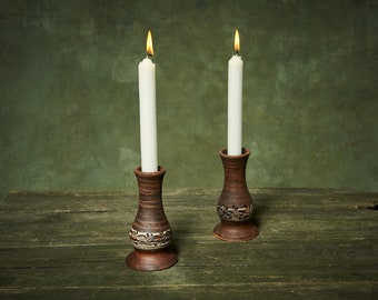 May include: Two lit white candles in brown ceramic candle holders with floral designs. The candle holders are on a weathered wooden surface against a green background. The flames are bright yellow.