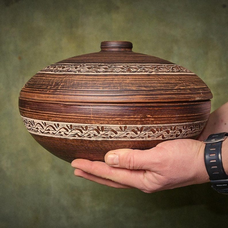 May include: A round, dark brown clay lidded bowl featuring horizontal striations and a decorative band of carved, light-colored leaf patterns encircling both the lid and the main body of the vessel, creating a textured, earthy appearance.