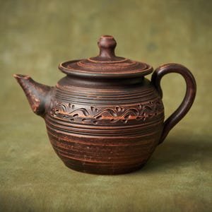 May include: A handcrafted brown ceramic teapot with a lid and handle. The teapot features a decorative floral pattern around the middle and is set against a green background. The teapot is a rustic style and is suitable for serving tea.