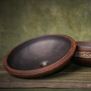 May include: A round, shallow ceramic bowl with a smooth, dark charcoal-colored interior and a reddish-brown rim. A second, partially visible bowl sits to the right, featuring a decorative band with a light-colored, intricate floral pattern.