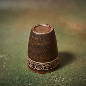 May include: A handmade, dark brown ceramic cup with a decorative band near the base. The cup has a textured surface and a slightly flared shape. The band features a carved, repeating pattern. The cup is displayed against a mottled green and brown background.