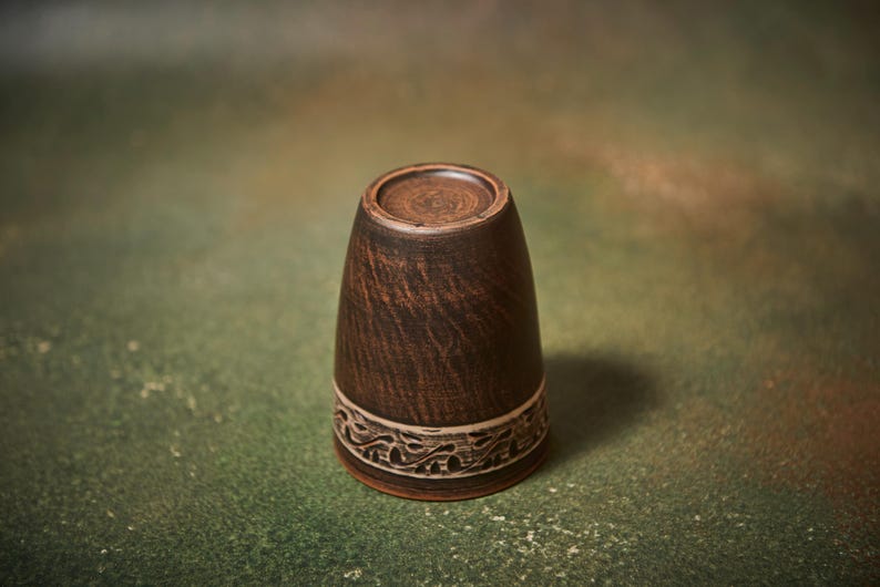 May include: A handmade, dark brown ceramic cup with a decorative band near the base. The cup has a textured surface and a slightly flared shape. The band features a carved, repeating pattern. The cup is displayed against a mottled green and brown background.