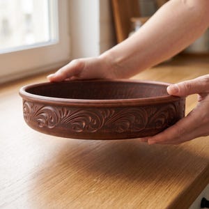 May include: A round, shallow terracotta bowl with a dark reddish-brown finish. The exterior wall of the bowl features a continuous, carved relief pattern of swirling floral motifs and leaves encircling the entire circumference of the vessel.