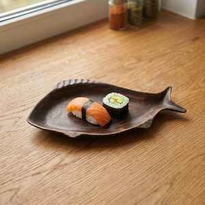May include: A brown, fish-shaped wooden plate holds two pieces of sushi: salmon nigiri and a cucumber roll. The plate has a textured surface and is placed on a wooden table near a window.