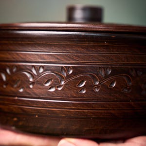 May include: A dark brown ceramic lidded bowl featuring a horizontal band of carved leaf patterns around the center. The bowl has a smooth, polished finish with subtle horizontal lines etched into the clay above and below the decorative vine motif.