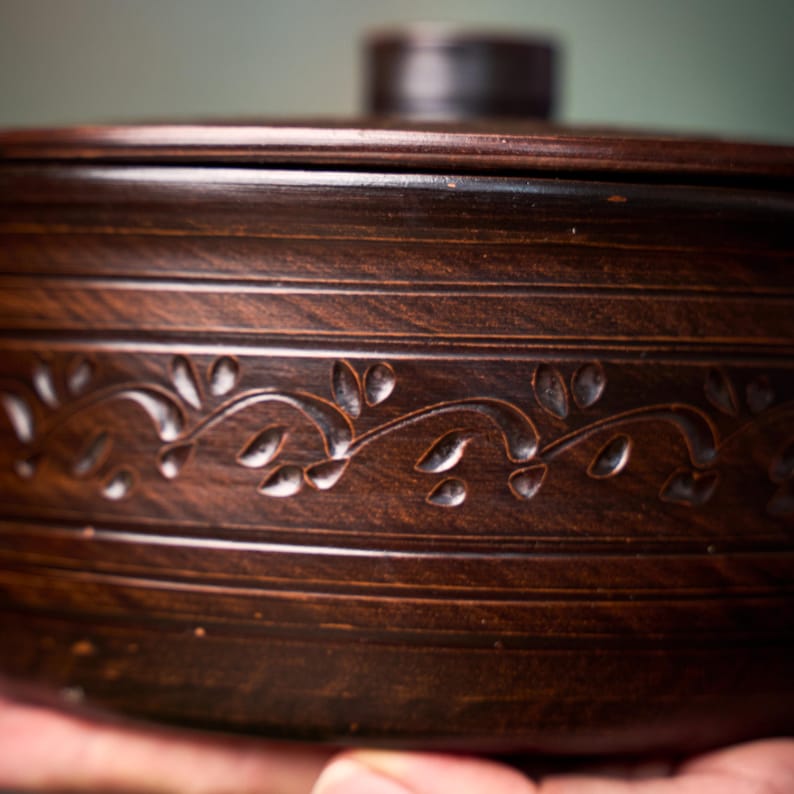 May include: A dark brown ceramic lidded bowl featuring a horizontal band of carved leaf patterns around the center. The bowl has a smooth, polished finish with subtle horizontal lines etched into the clay above and below the decorative vine motif.