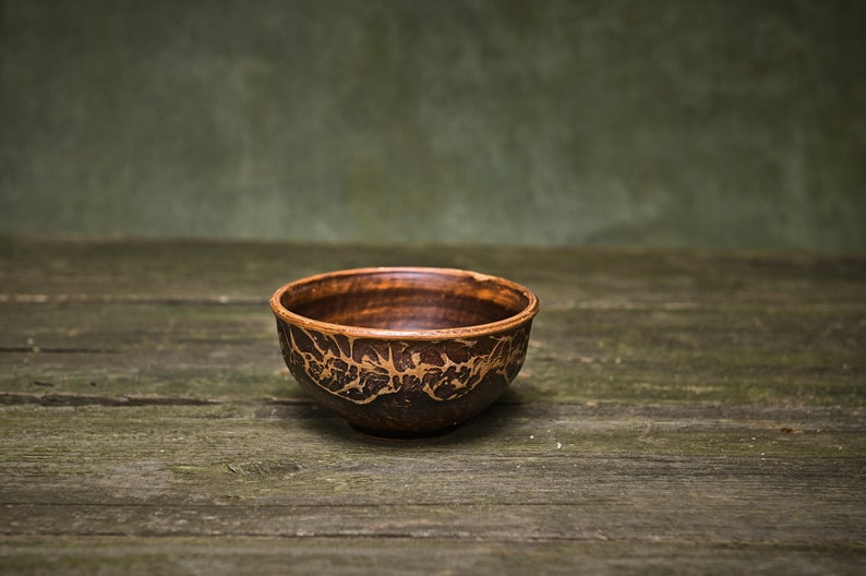 May include: A small, brown ceramic bowl with a decorative pattern. The bowl is sitting on a rustic wooden surface.