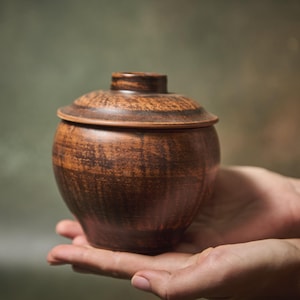 Peut inclure: Un petit pot rond en argile brune avec un couvercle assorti. Le récipient présente une surface texturée et marbrée avec des stries brun foncé, offrant une finition naturelle et terreuse ainsi qu'un design simple et fonctionnel.