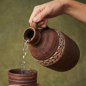 Könnte beinhalten: Ein brauner Keramikkrug mit Henkel gießt Wasser in eine passende Tasse. Der Krug hat ein dekoratives Band in der Mitte. Der Hintergrund ist ein gedämpftes Grün.