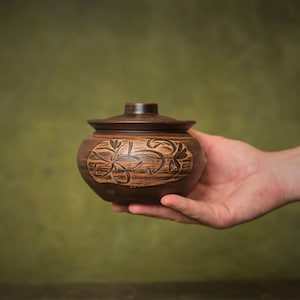 Op de afbeelding: Een kleine bruine keramische pot met een deksel, met een gegraveerd bloemenmotief. De pot wordt in de hand van een persoon gehouden.