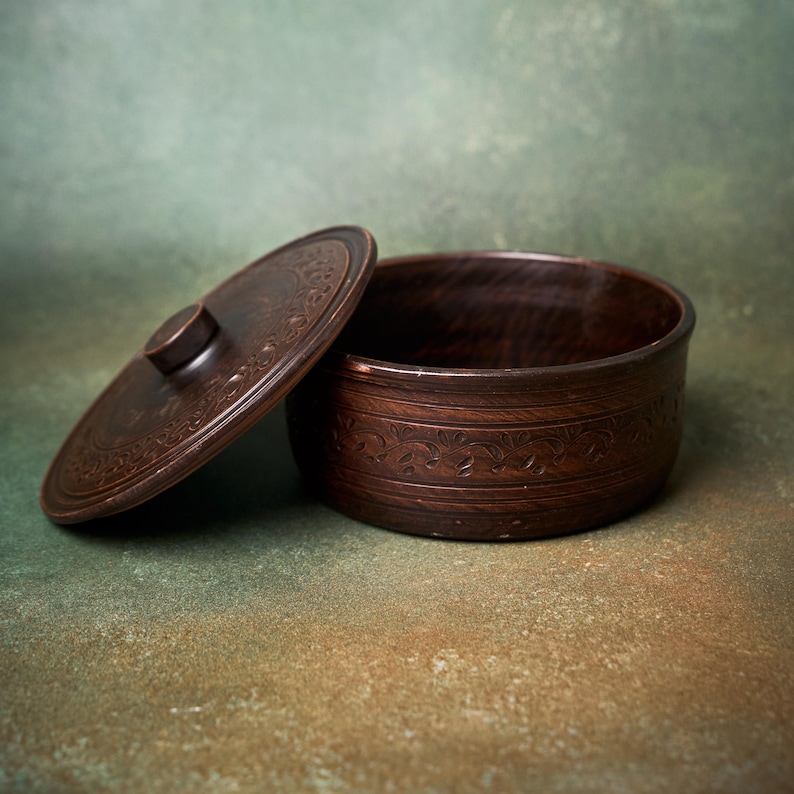 May include: A round, dark brown clay container with a matching lid. The exterior of the bowl and the top of the lid feature a carved, repeating floral vine pattern. The lid has a small, centered circular knob for lifting.