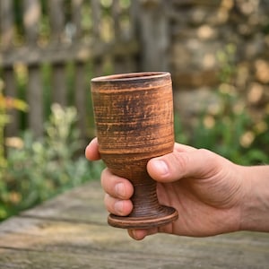 May include: A brown clay goblet with a textured, rustic finish. The vessel features a cylindrical bowl that tapers down to a narrow stem, ending in a wide, circular base. The surface displays natural variations in color, ranging from light tan to dark brown.