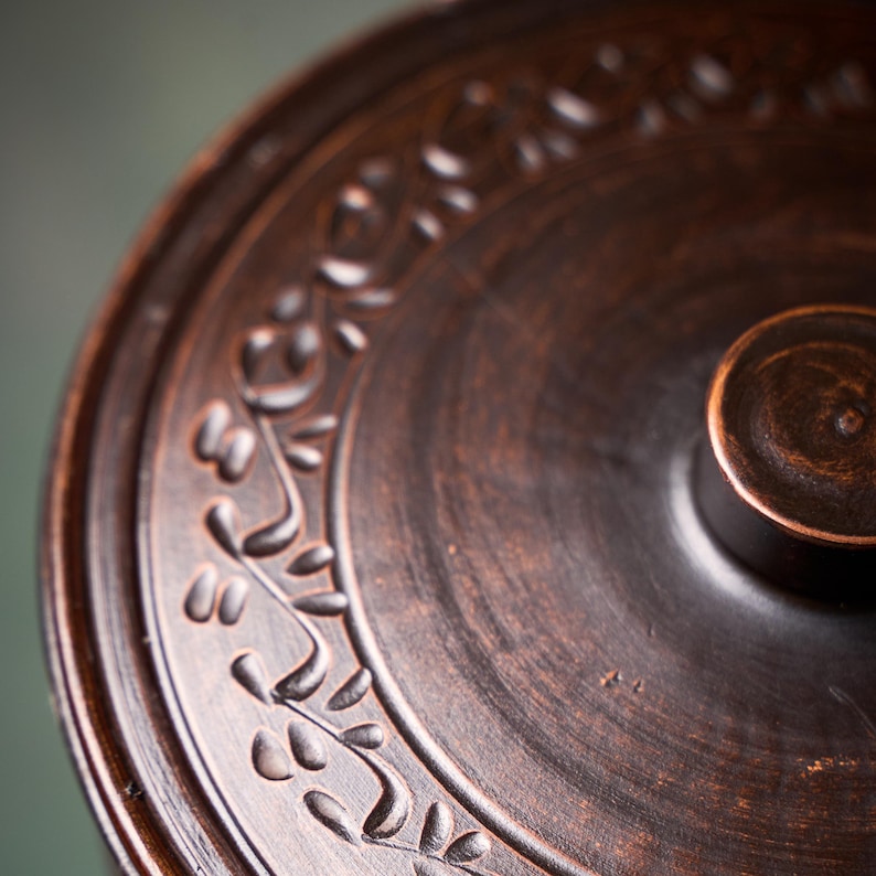 May include: A circular dark brown clay lid with a smooth, polished finish and a central knob handle. The surface features a decorative embossed pattern of small, stylized leaves arranged in a continuous border along the outer edge of the lid.