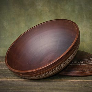 May include: A deep, round, dark brown clay bowl with a smooth interior surface and a wide, flat rim. The exterior side of the bowl features a decorative, repeating carved pattern that encircles the circumference of the vessel near the base.