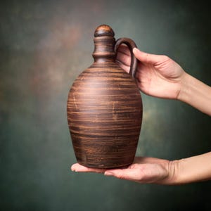 May include: A brown clay jug with a rounded body, a narrow neck, and a single handle on the side. The surface features horizontal striations and a smooth, dark brown stopper at the top. The vessel has a rustic, earthy appearance.