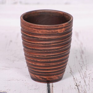May include: A cylindrical brown clay cup featuring a series of horizontal ridges along the exterior surface. The cup has a dark, earthy tone with subtle variations in color and a textured finish that highlights the circular grooved pattern.
