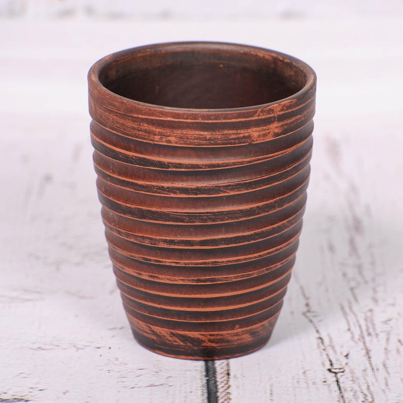 May include: A cylindrical brown clay cup featuring a series of horizontal ridges along the exterior surface. The cup has a dark, earthy tone with subtle variations in color and a textured finish that highlights the circular grooved pattern.