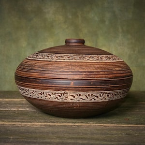 May include: A round, brown clay lidded bowl featuring horizontal ridges and two bands of carved, light-colored leaf patterns. The bowl has a smooth, dark brown finish with visible texture and a matching lid with a small, centered knob handle.