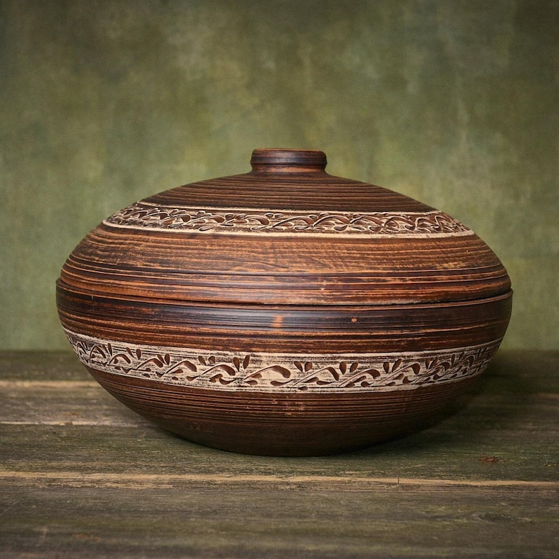 May include: A round, brown clay lidded bowl featuring horizontal ridges and two bands of carved, light-colored leaf patterns. The bowl has a smooth, dark brown finish with visible texture and a matching lid with a small, centered knob handle.