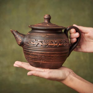 May include: A dark brown clay teapot featuring a rounded body, a matching lid with a small knob, a curved handle, and a short spout. The exterior surface displays horizontal ridges and a decorative band of carved leaf-like patterns around the center.