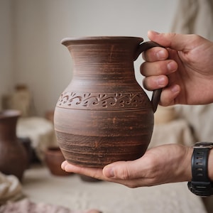 Peut inclure: Une cruche en argile brune avec un corps arrondi et un bord évasé. La surface présente des stries horizontales et une bande décorative de motifs en forme de feuilles sculptées sur la partie supérieure. Elle comprend une anse incurvée.