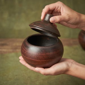 May include: A round, dark brown clay pot with a matching lid. The pot has a smooth, slightly textured surface with visible horizontal striations in the clay. The lid features a small, central knob for lifting, and the pot has a wide opening.