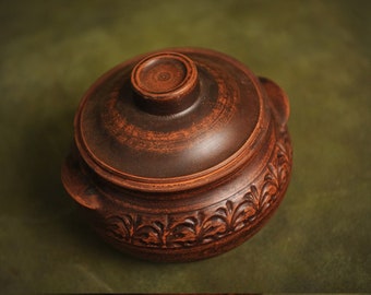 May include: A handcrafted, brown ceramic pot with a lid. The pot features a decorative floral pattern around the base and a small handle on the lid. The pot is likely used for cooking or serving food.