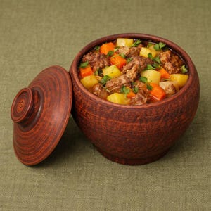 Puede incluir: Una rústica olla de cerámica marrón llena de un guiso abundante. El guiso contiene trozos de carne, zanahorias naranjas y patatas amarillas, adornado con hierbas verdes frescas. La tapa está colocada al lado sobre una tela verde.