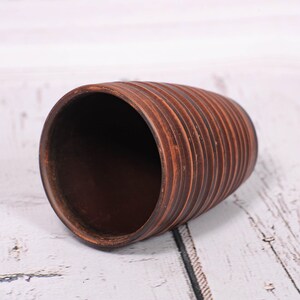 May include: A cylindrical brown clay cup featuring a series of horizontal, raised ridges encircling the exterior. The interior of the cup is smooth and dark brown, while the outer surface displays a textured, ribbed pattern in earthy tones.