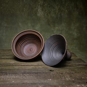 May include: Two dark ceramic bowls with a distinct circular ridged texture on the interior surfaces. One bowl is positioned upright showing a reddish-brown hue, while the other is tilted on its side, displaying a matte black finish and a small handle.