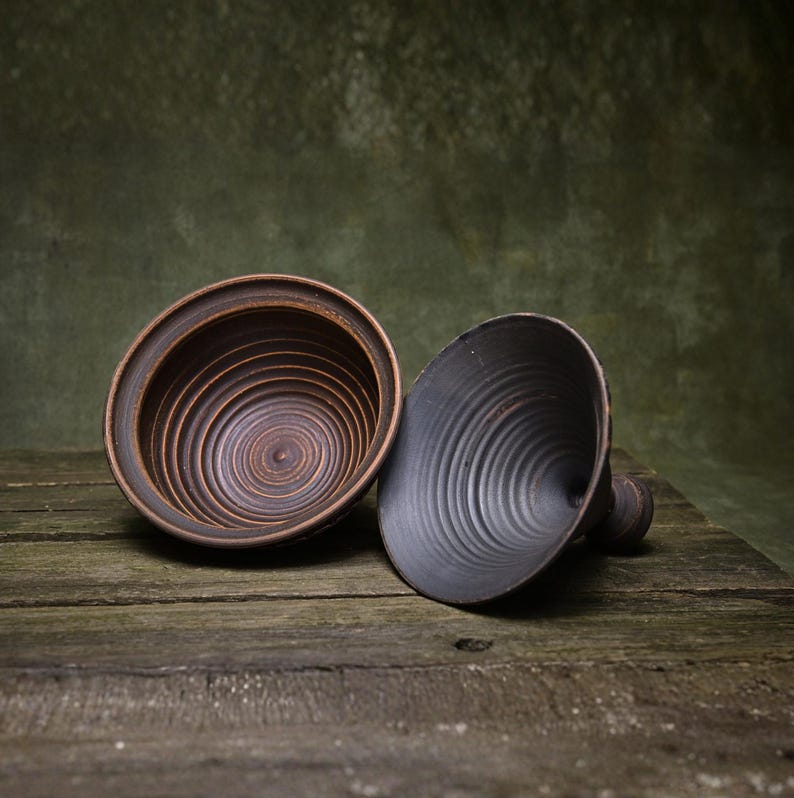 May include: Two dark ceramic bowls with a distinct circular ridged texture on the interior surfaces. One bowl is positioned upright showing a reddish-brown hue, while the other is tilted on its side, displaying a matte black finish and a small handle.
