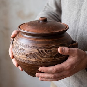 May include: A round, dark brown clay pot with a matching lid and two side handles. The exterior features a carved, horizontal wave-like pattern encircling the middle section, showcasing the natural texture and earthy tones of the fired ceramic material.